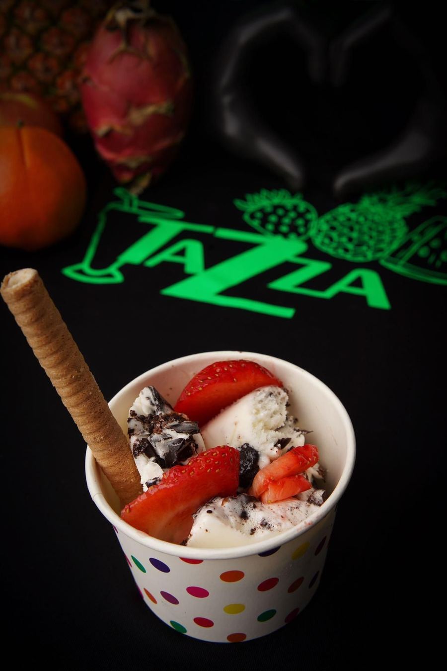 A cup of Tazza gelato with strawberries and a wafer roll, lit against the shop's green neon Tazza sign.