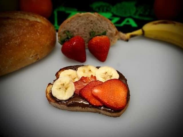 Nutella toast topped with sliced banana and strawberry, with a loaf and whole fruit behind.