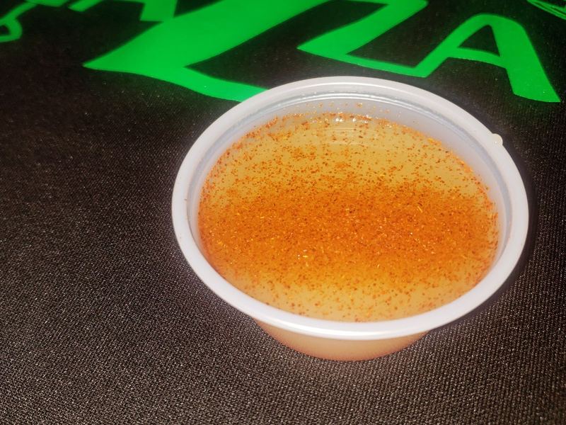 A small cup of golden-orange immune shot flecked with spice, on a dark counter under the green Tazza sign.