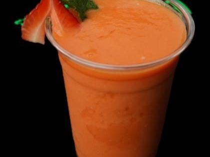 A tall clear cup of bright orange smoothie garnished with a strawberry and a sprig of mint.