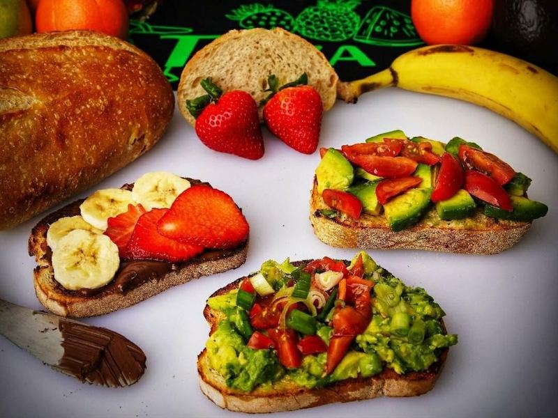 Three slices of Tazza toast — two avocado, one Nutella with banana and strawberry — with a loaf and fresh fruit behind them.